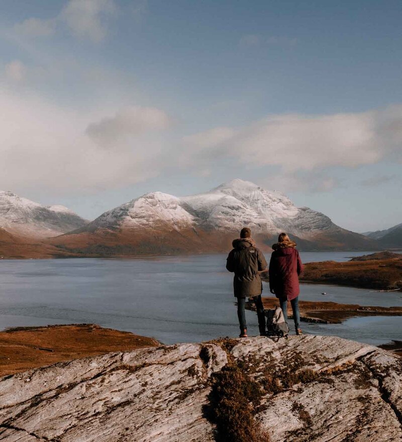 Photo prise par drone de Deux Drôles d’Oiseaux, de dos au sommet d’une montagne face à un lac dans les Highlands en Écosse – un instant suspendu entre nature sauvage et complicité silencieuse