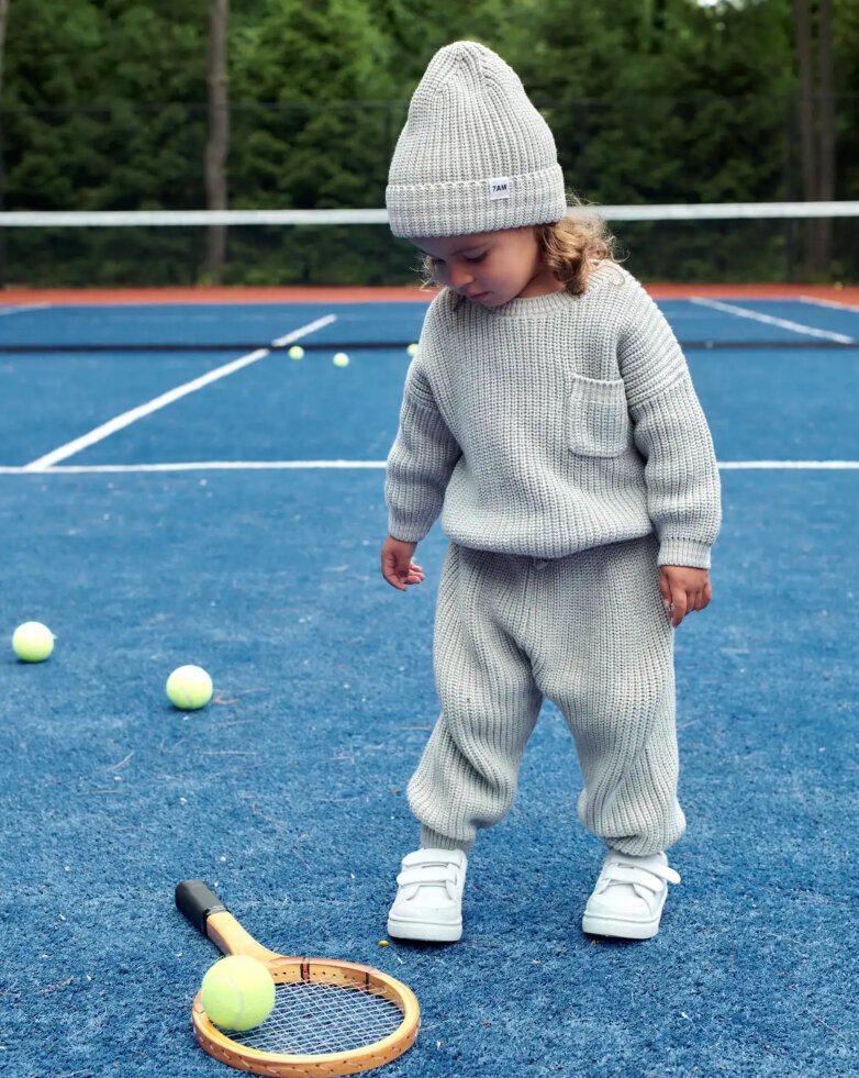 A baby looking at a tennis racket and tennis ball on the ground.