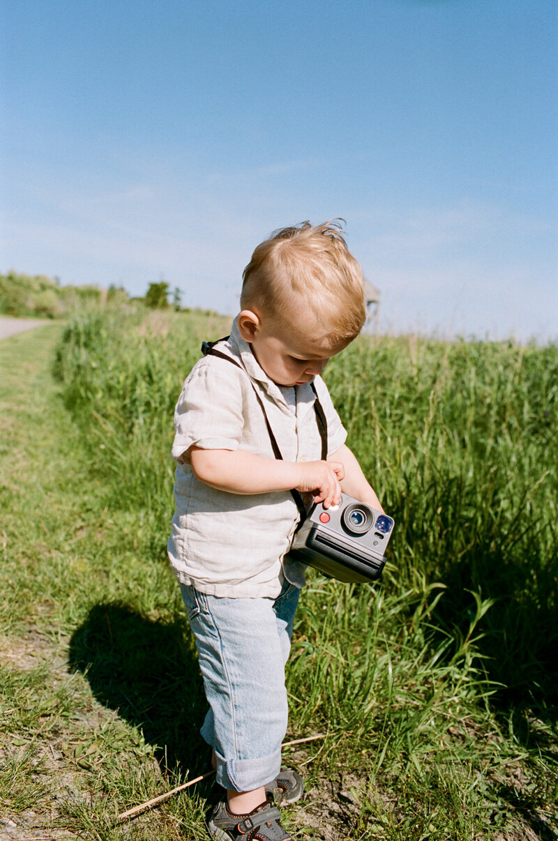 A child stands in a sunlit field, holding a vintage camera, with grass swaying against a clear blue sky—capturing moments through the lens of family photography Toronto cherishes.
