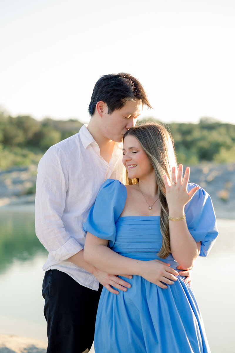 Romantic proposal at Pedernales Falls State Park — woman in blue dress showing engagement ring while her partner embraces her in golden hour light.