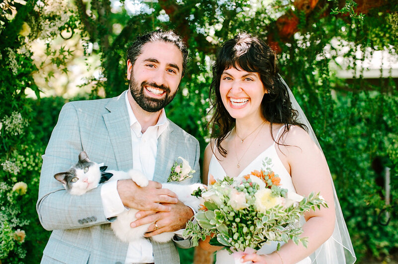 Bride and groom smiling outdoors, holding a bouquet and their cat.