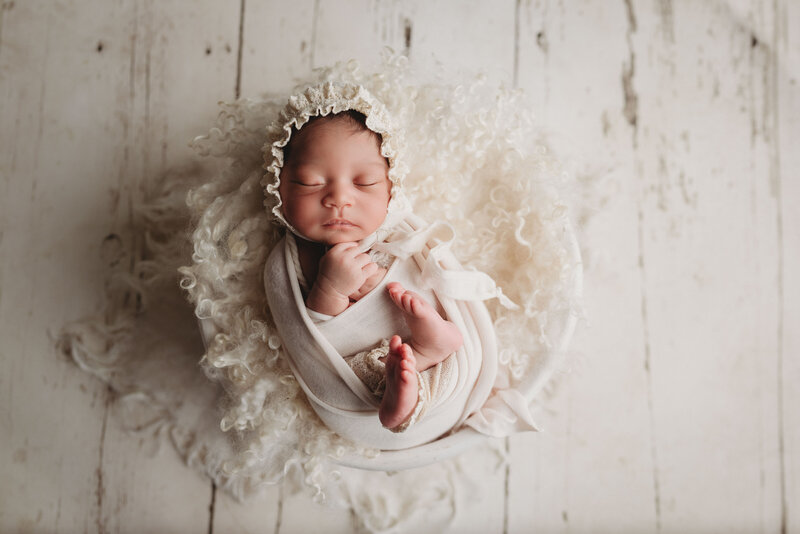 Beautiful newborn baby girl wrapped and swaddled in cream color wrap, bonnet and fur laying in white bowl on white wood floors