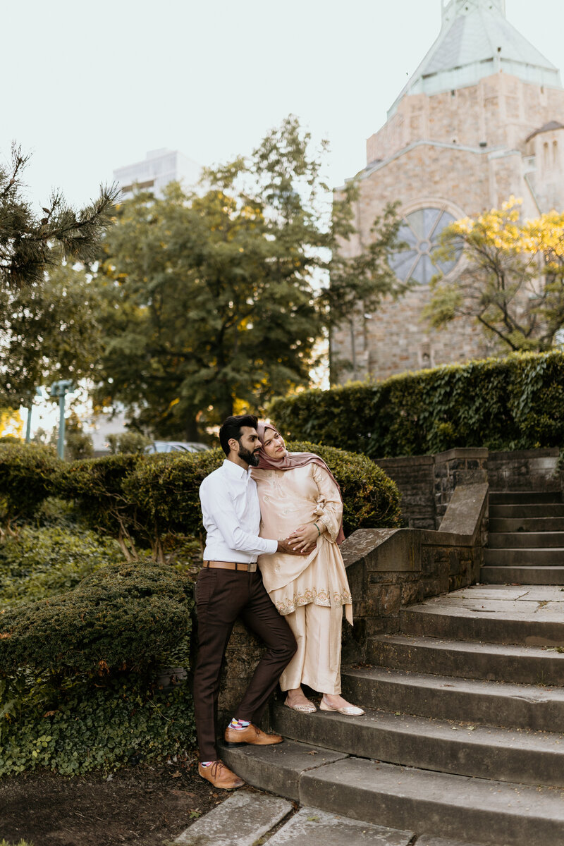 A husband and his pregnant wife embrace outside some steps in the city