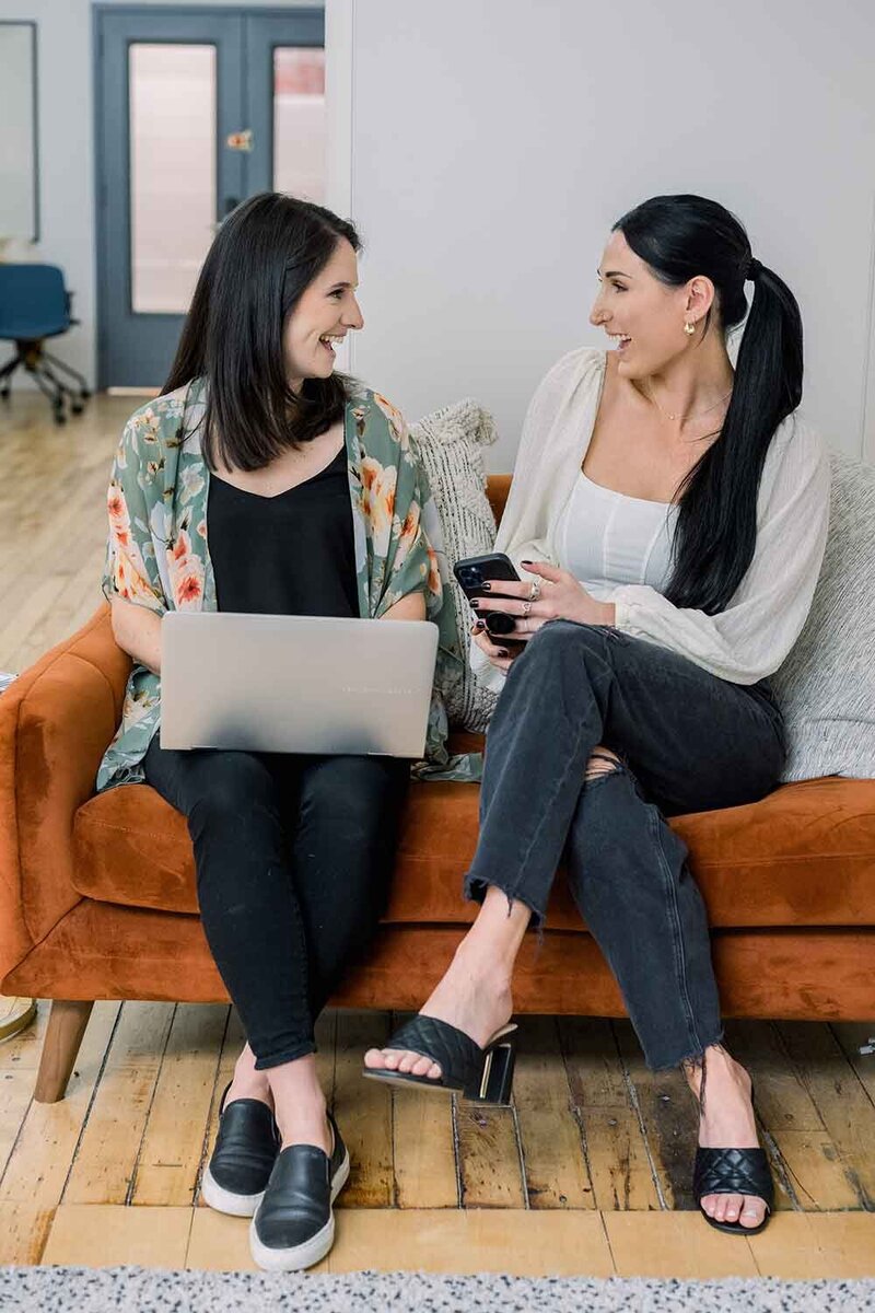 Two women sitting on couch, smiling at each other,, one has a phone in her hand and the other has her laptop