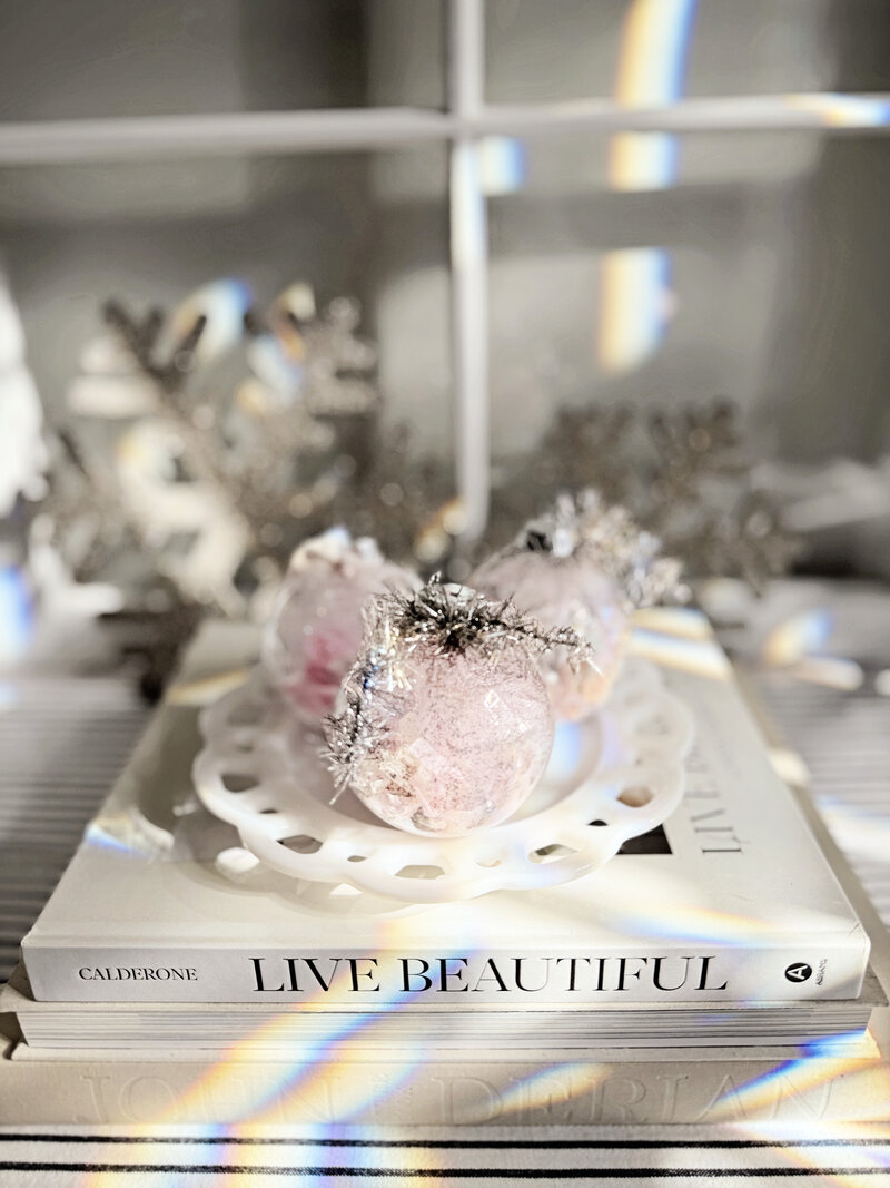 Three pink holiday ornaments with silver tinsel sit on a white dish atop stacked books, including one titled "Live Beautiful," with rainbow light reflections across the scene—a charming display by Sweet Pea Ink Creative.
