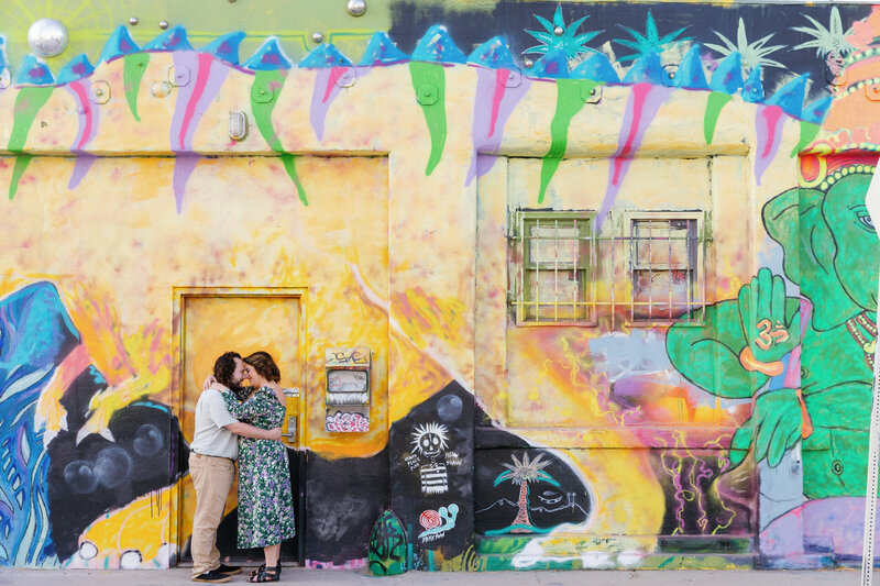 A couple with their arms around each other standing in front of a brightly colored mural