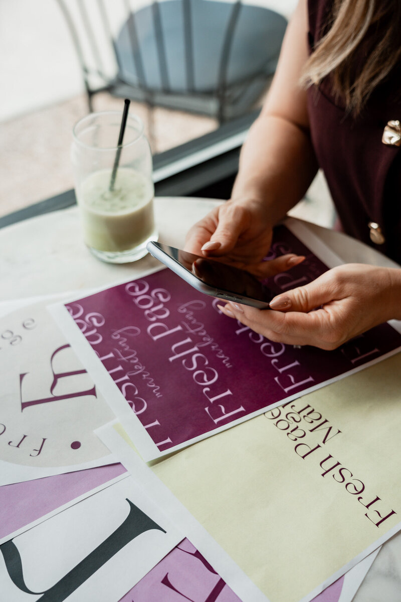 Hands holding smartphone over Fresh Page Marketing brand papers, showcasing digital marketing support for Shopify, Faire, copywriting, and web design.