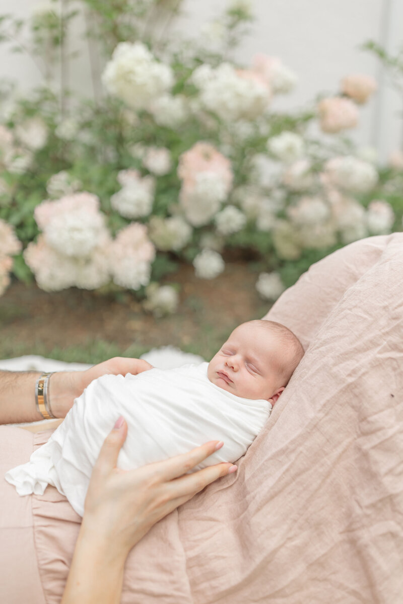 A mother lies on her back in a garden, gently cradling her newborn on her legs. Surrounded by lush greenery and blooming flowers, she gazes lovingly at her baby, who rests peacefully. Sunlight filters through the leaves, casting a warm, natural glow over the tender moment