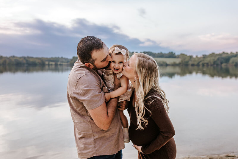 Expecting parents kiss their smiling toddler during a lakeside family maternity session at sunset.