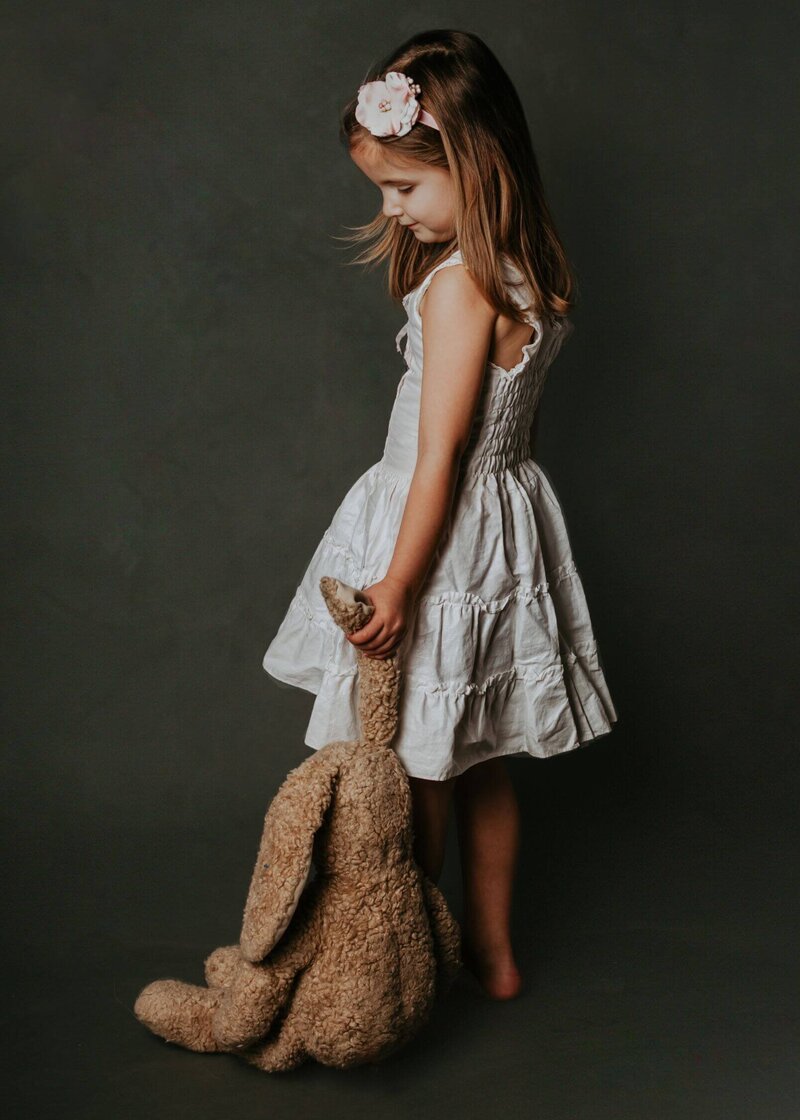 A little girl in a plain white dress hold her stuffed bunny by the ear