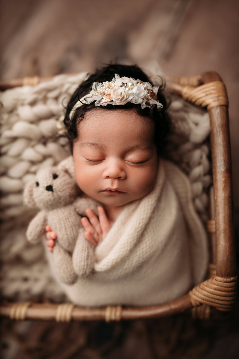 2 week old newborn baby girl wrapped in a cream textured swaddle wearing a floral headband and holding teddy bear inside of a brown basket