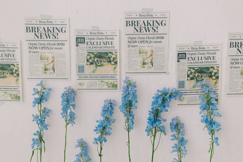 Editorial floral announcement wall featuring blue flowers and printed styled newspaper clippings for a 2026 event launch. Clean white background with creative flat lay design. Greenville South Carolina portrait photographer – Dani Lacey Photograph
