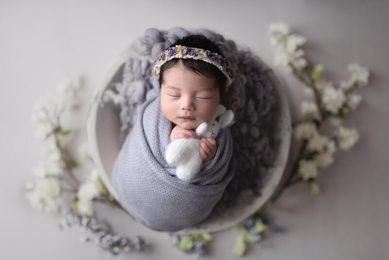 Newborn girl in lavender wrap with floral crown, holding white bunny plush in soft spring-inspired studio setup