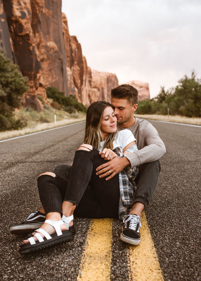Couple sitting in the middle of a scenic desert road embraced by red rock cliffs, captured by a Colorado Wedding and Portrait Photographer.