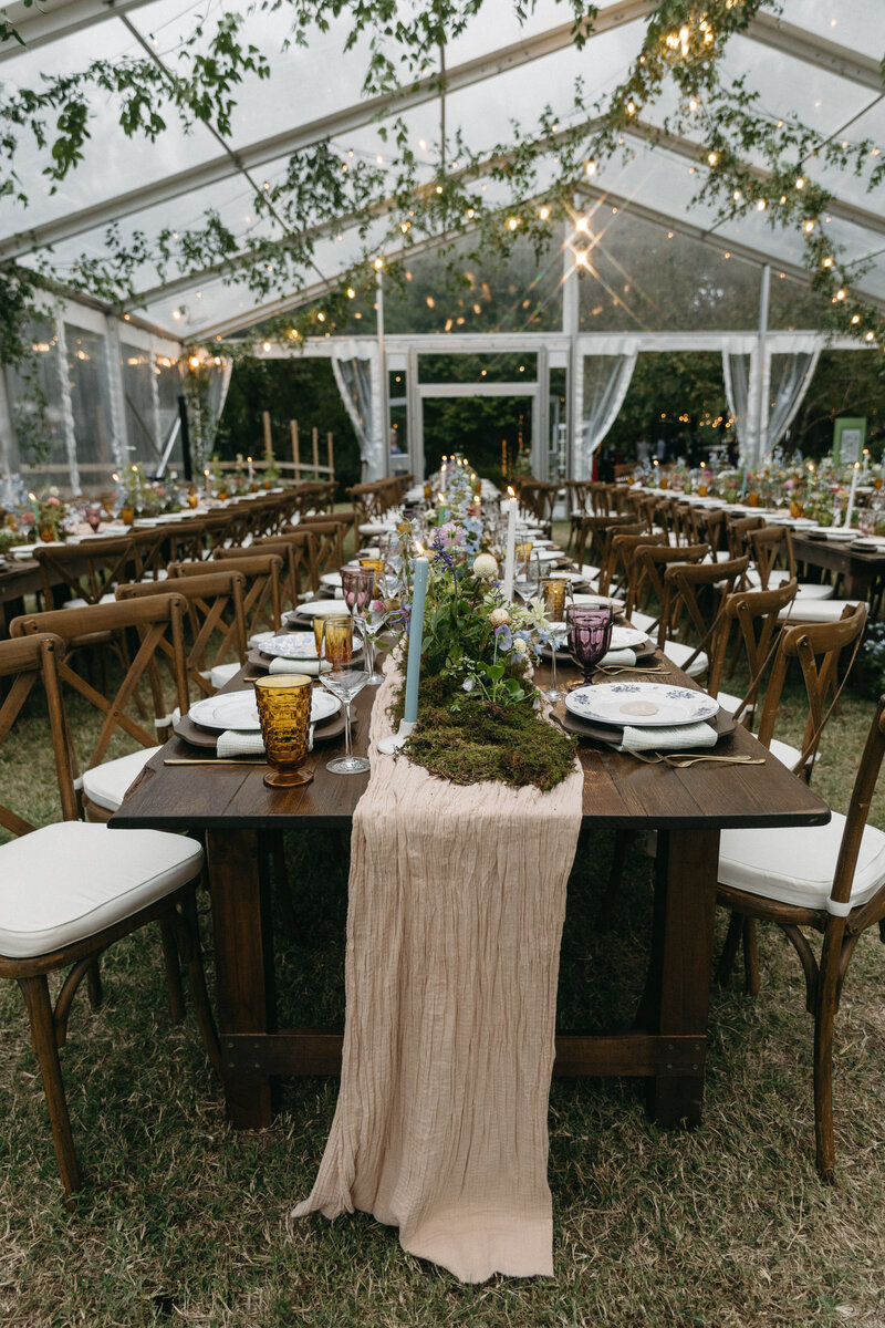 A lush garden-inspired wedding reception tablescape set beneath a clear tent, featuring long wooden farm tables decorated with a soft blush table runner, natural moss accents, pastel taper candles, vintage mismatched glassware, and whimsical wildflower centerpieces with blue, purple, and yellow blooms, creating an elegant outdoor celebration atmosphere.