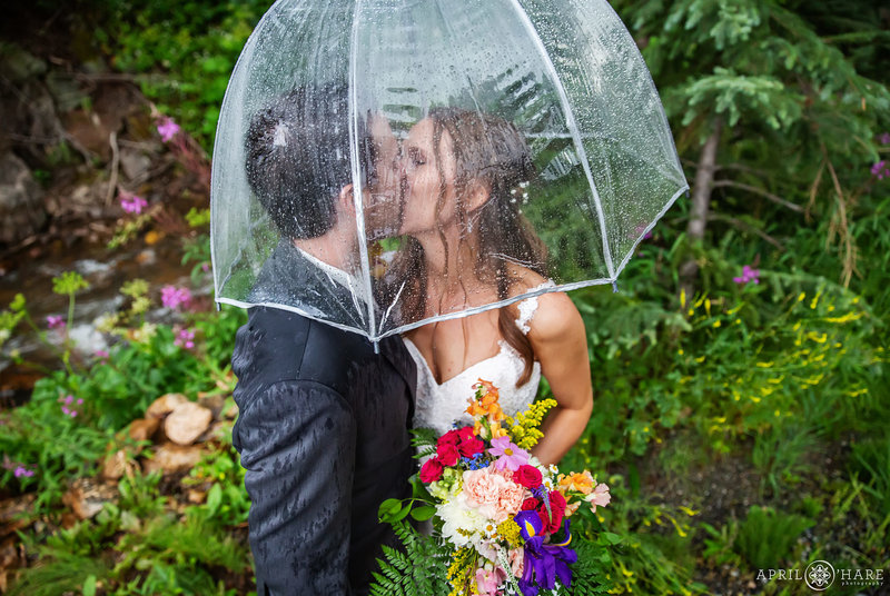 Couple kiss on their outdoor rainy wedding day under a clear umbrella in the garden at Blackstone Rivers Ranch