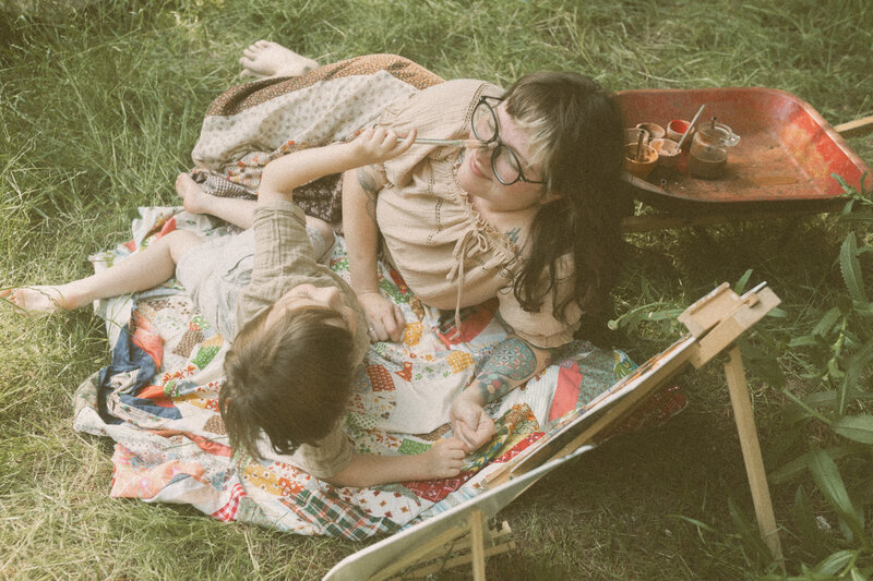 Two sisters lay on a quilted blanket together while the youngest pokes the older sisters nose. 
