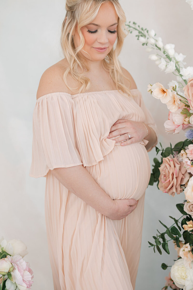 Pregnant mom in a light pink dress standing next to pink flowers as she holds her baby bump and looks down at it with a soft smile