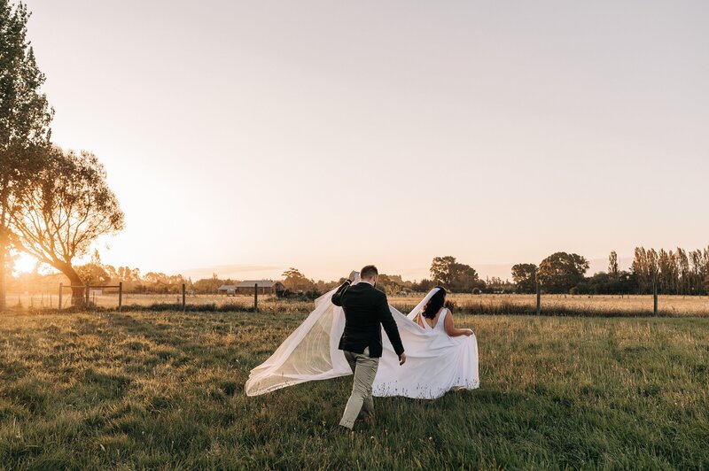 loving ellies belly wedding sunset groom holding brides train christchurch nz walking across field