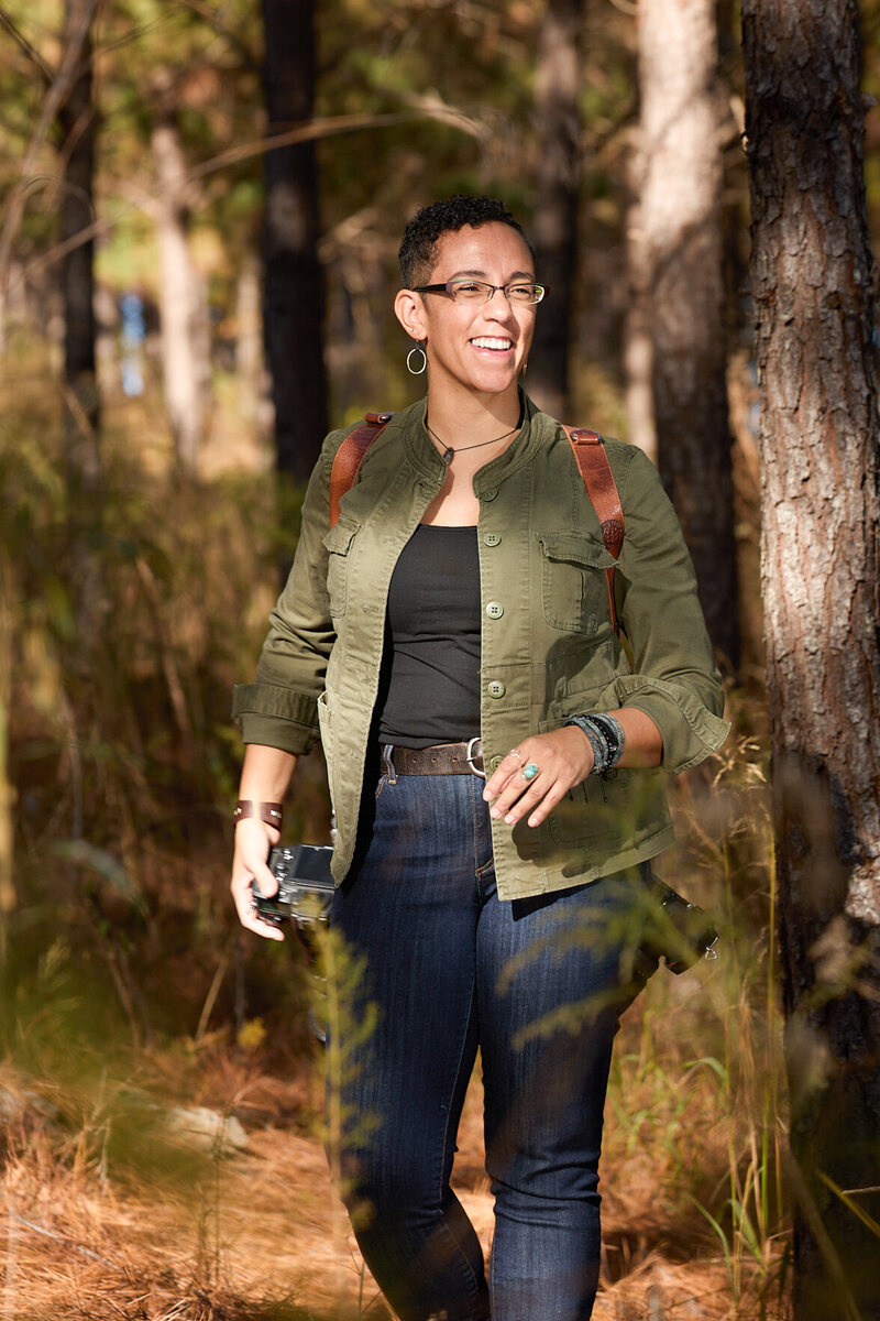 image of Kate Okenatez-Mahoney, a top photographer for destination elopements and intimate weddings.  She has curly, short black hair, medium brown skin, and is wearing a safari inspired olive green jacket, blue jeans, and carrying two big cameras on a leather holster. she has on turquoise rings, and modern-styled eyeglasses with a big smile looking away from the camera.