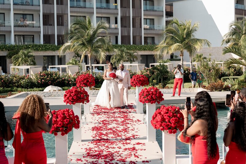 A bride and groom walk down a dock with red rose petals.