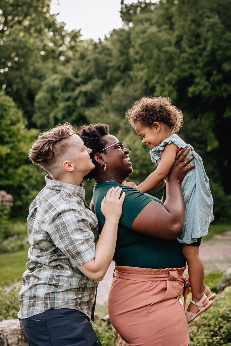 A joyful family scene where two parents hold their toddler between them. The toddler, in a light blue dress, is being lifted by one parent while all three share a warm, affectionate moment outdoors.