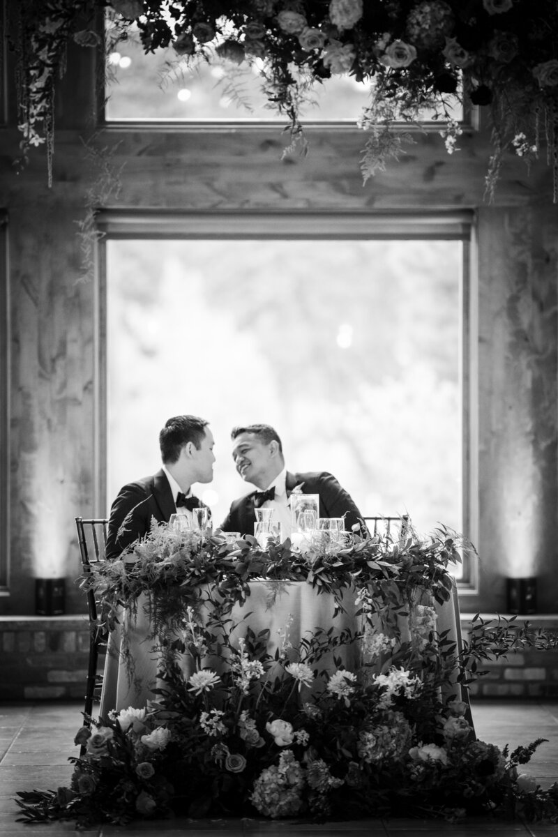 Two groom sitting at a wedding reception table about to kiss.