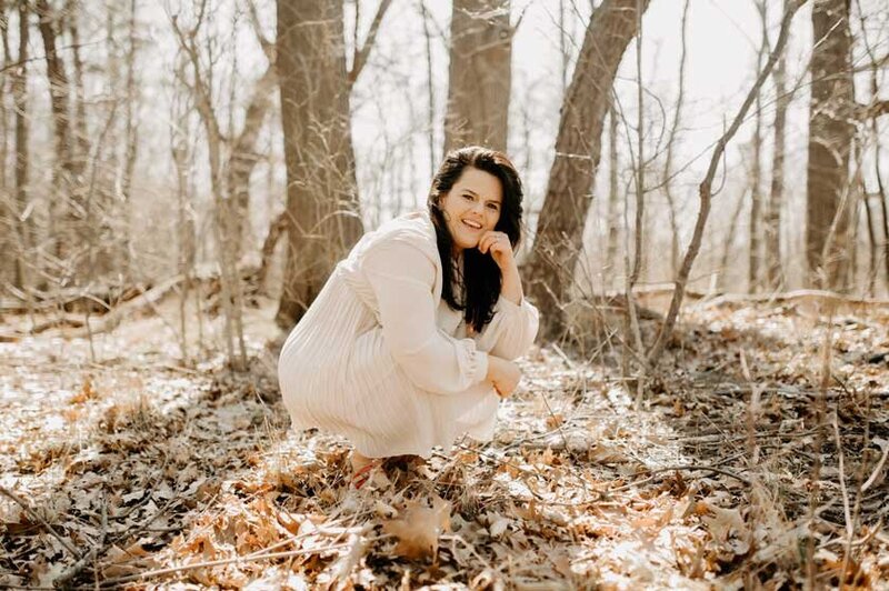 Kathleen Oh in a beige dress crouched down in the woods with her hand on her chin.