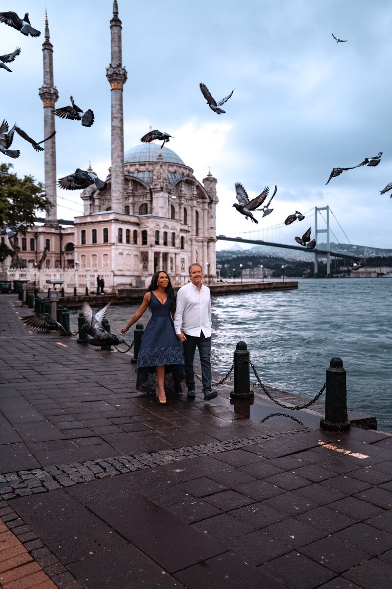 Couple walks along the water with birds flying overhead