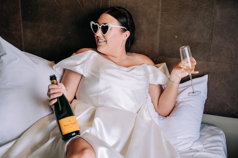 a bride in paperswan dress with bottle of veuve and heart sunglasses in bed