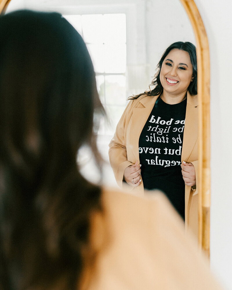 A woman smiles at her reflection in a mirror, wearing a tan blazer over a black shirt with white text. The room is bright, conveying a positive atmosphere.
