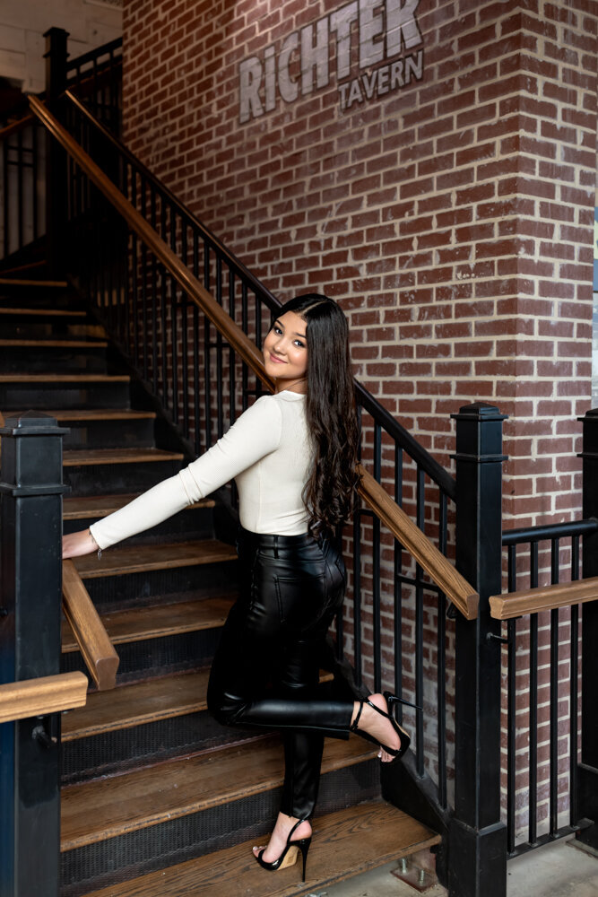 teele High School senior girl standing on stairs with one leg kicked back wearing black pants and white shirt during session in Boerne, Texas.