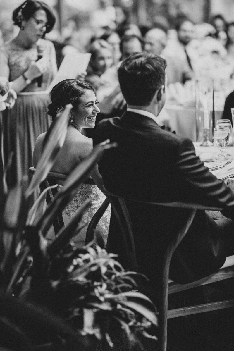 black and white of white groom in black tux and white brunette bride in strapless white gown smiling at one another sitting with their backs to the camera at their sweetheart table while parents are giving a speech