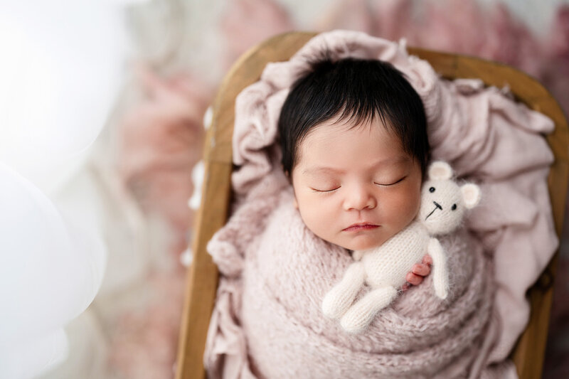 Newborn baby girl with lots of long, dark hair sleeps in pink knit swaddle while holding tiny pink teddy bear in Jen Howell Photography's Memphis newborn photography studio.