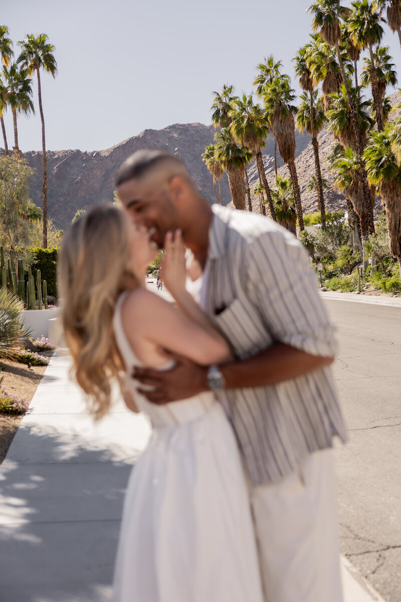 A dreamy and romantic moment captured in the heart of Palm Springs, California. With a soft, artistic blur, this intimate embrace between a couple highlights the warmth of love against a stunning desert backdrop of palm trees and mountains.