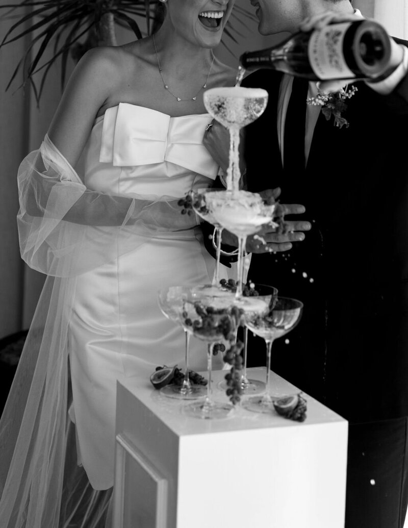A bride and groom share a joyful moment pouring champagne into a tower of glasses, adorned with figs and grapes.