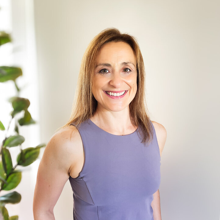 Vanessa Flores, a midlife woman coach in a lavender tank top smiles directly at the camera, standing in her home gym with soft natural light and gym equipment blurred in the background.