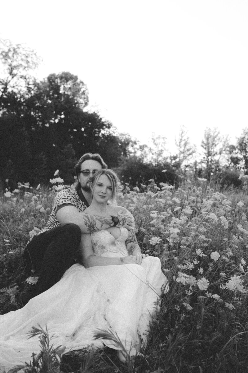 Bride and groom sitting amidst Queen’s lace flowers, looking at the camera in an editorial portrait style, captured by Emily Ambie.