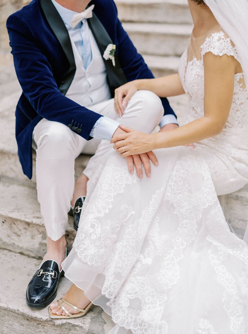 bride-groom-sit-old-stone-steps-place-their-hands-each-other-laps-cropped-faceless