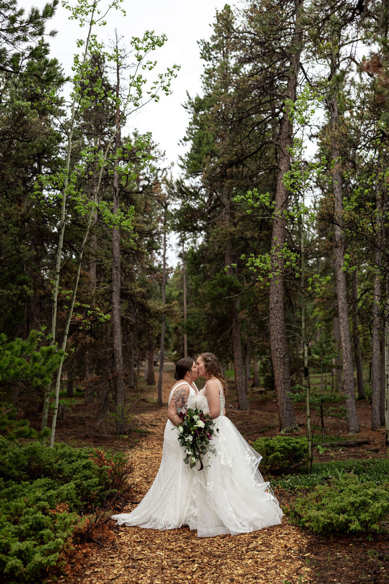 Two brides kissing on a small path in a forest.
