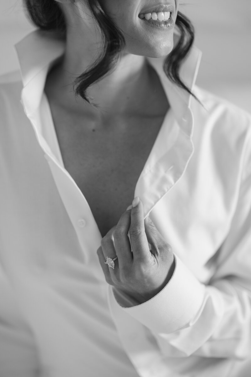 Black and white getting ready photo of bride wearing a white tuxedo shirt looking out the window by Suzanne Karp 