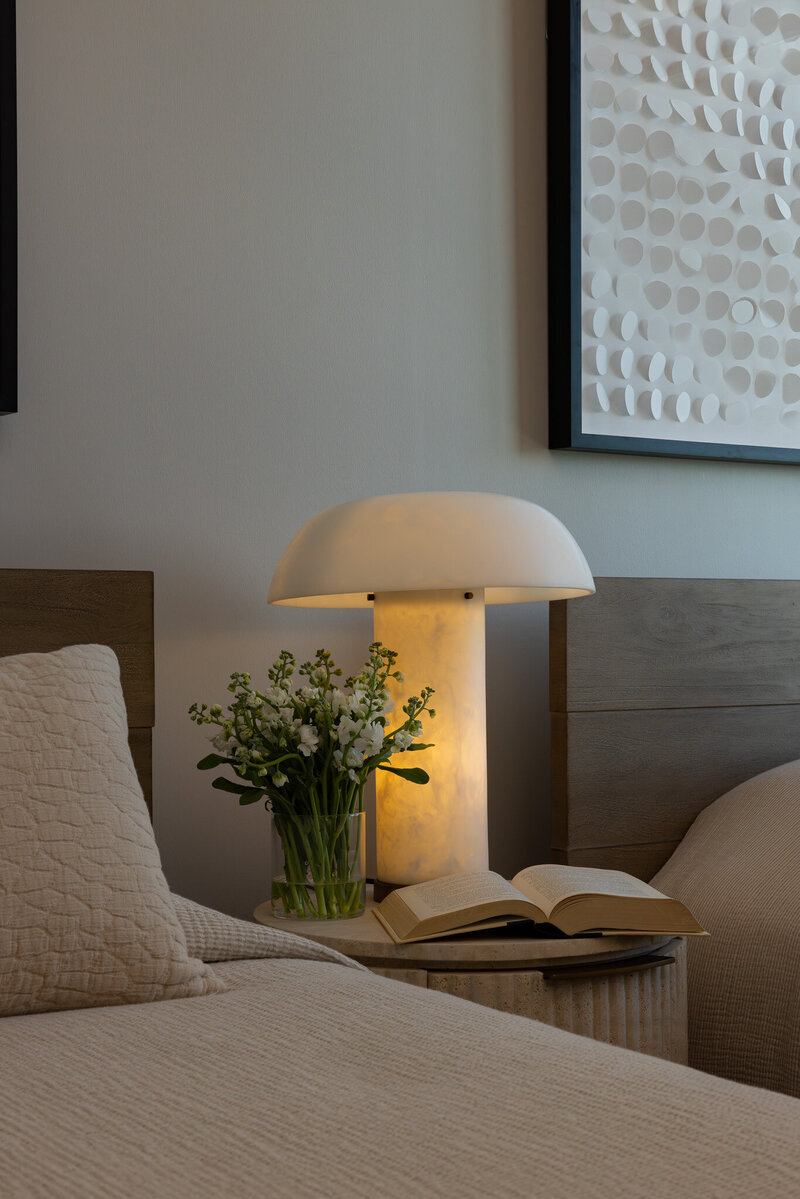 Detail of a secondary bedroom featuring warm wood bedframes, textured bedding, a fluted travertine nightstand with flowers and a book, designed by Sister Studio.