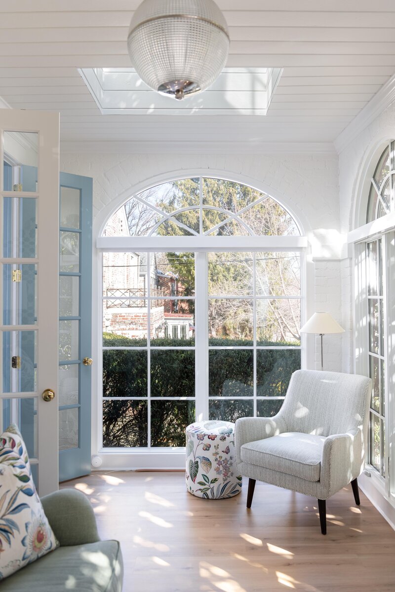 Room featuring arched windows, white armchairs, a round table, and floral cushions, bathed in natural sunlight