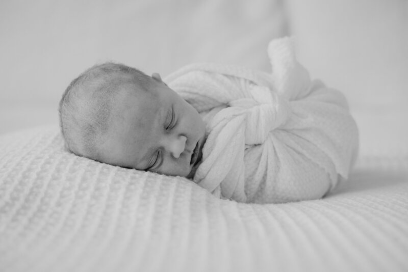 Black and white newborn photo of baby girl wrapped in blanket on a bed.
