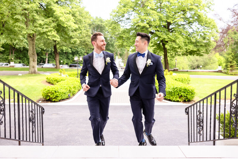 Two grooms joyfully holding hands as they ascend the steps on their wedding day, dressed in navy tuxedos with blue bow ties.