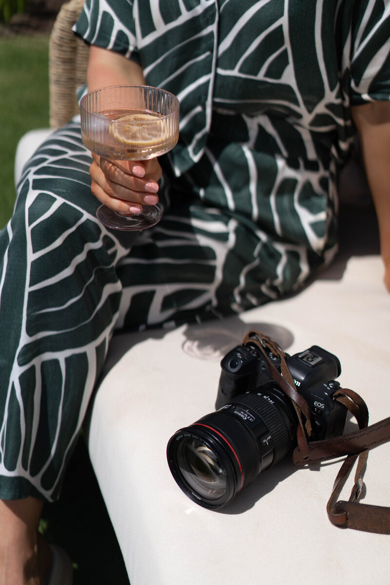 Jessie in a green pants outfit, cocktail in hand, with her camera by her side. Face not visible.

