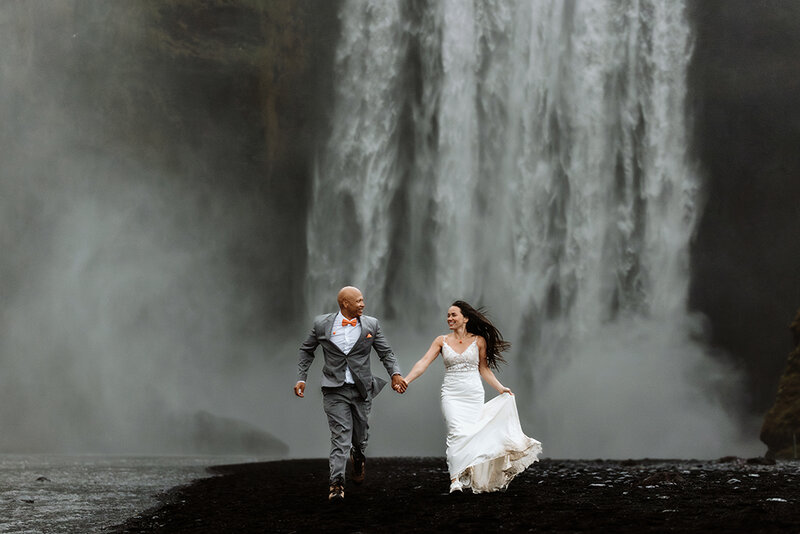 Bride and groom are laughing and running in front of the Skogafoss waterfall on their wedding day in Iceland