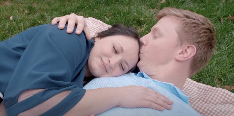 A couple laying in the grass together while one kisses the other's head 