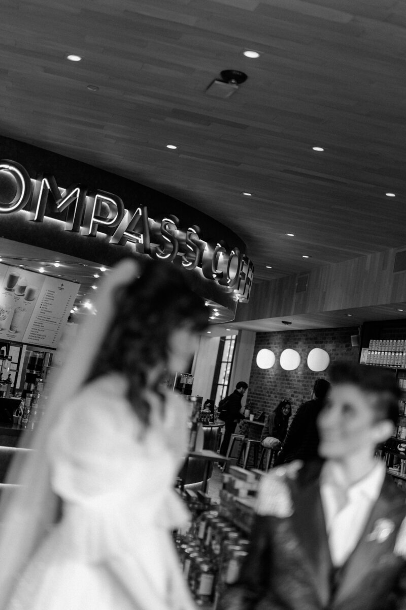 Black and white blurred image of out of focus nonbinary couple smiling at one another with the Compass Coffee Sign in focus at Compass Coffee in Georgetown DC 