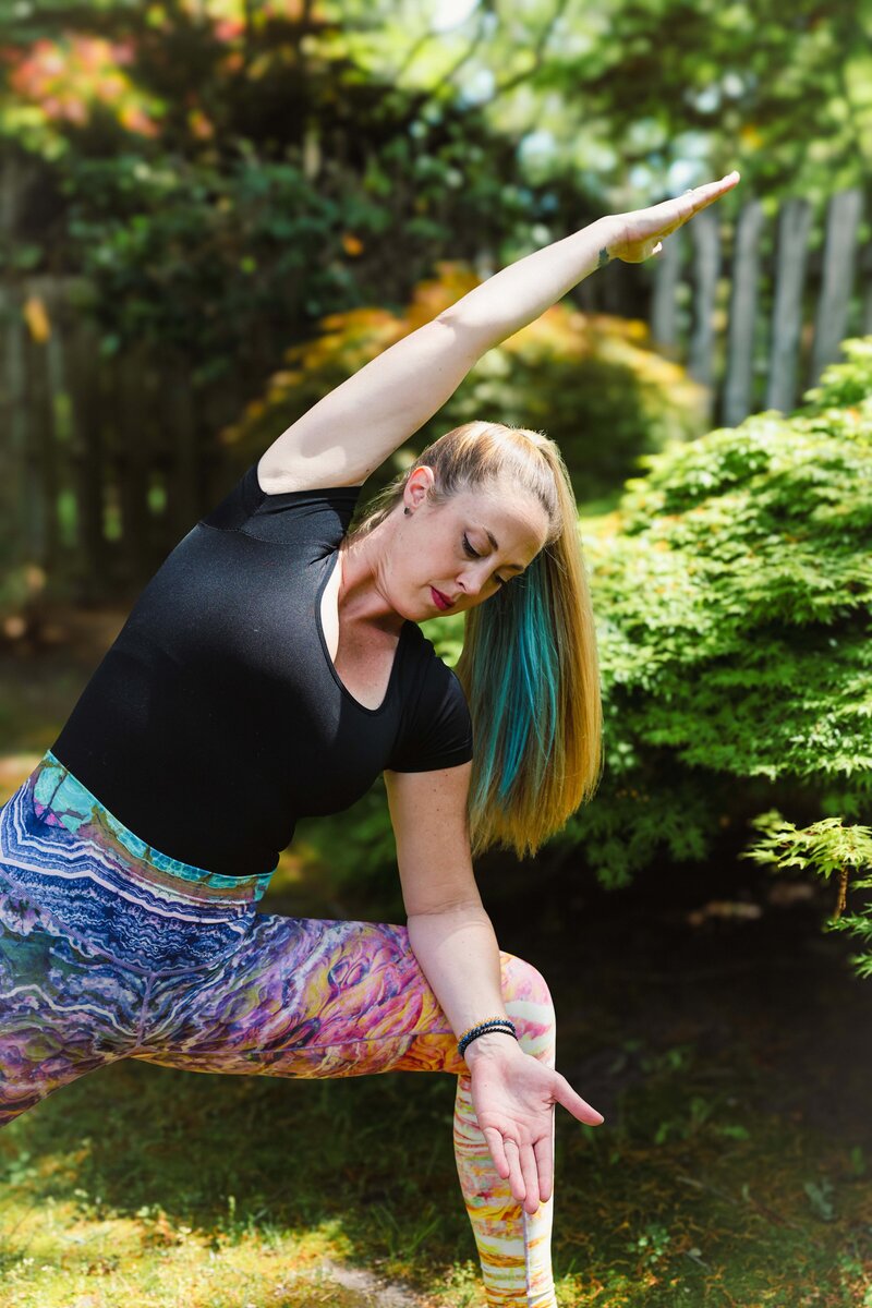 Woman wearing a black top and colorful yoga pants, standing in triangle pose in front of lush green trees in the background.